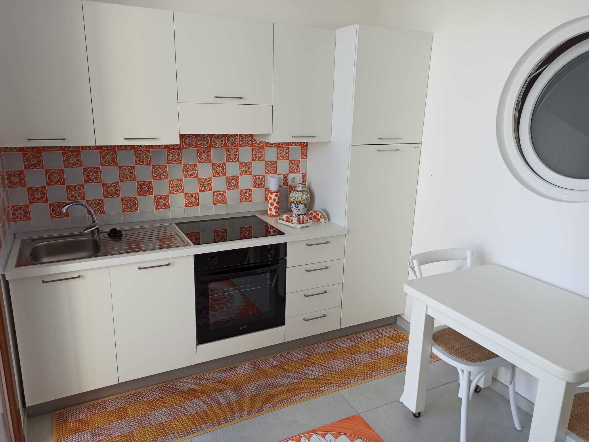 Uma cozinha compacta com armários brancos, forno embutido e uma pequena mesa de jantar com uma cadeira.