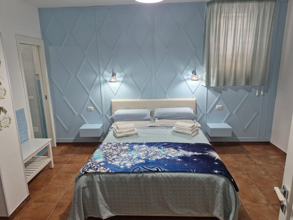 A bedroom with a bed, two bedside tables, a nightstand, a wall-mounted lamp, and a window with curtains.