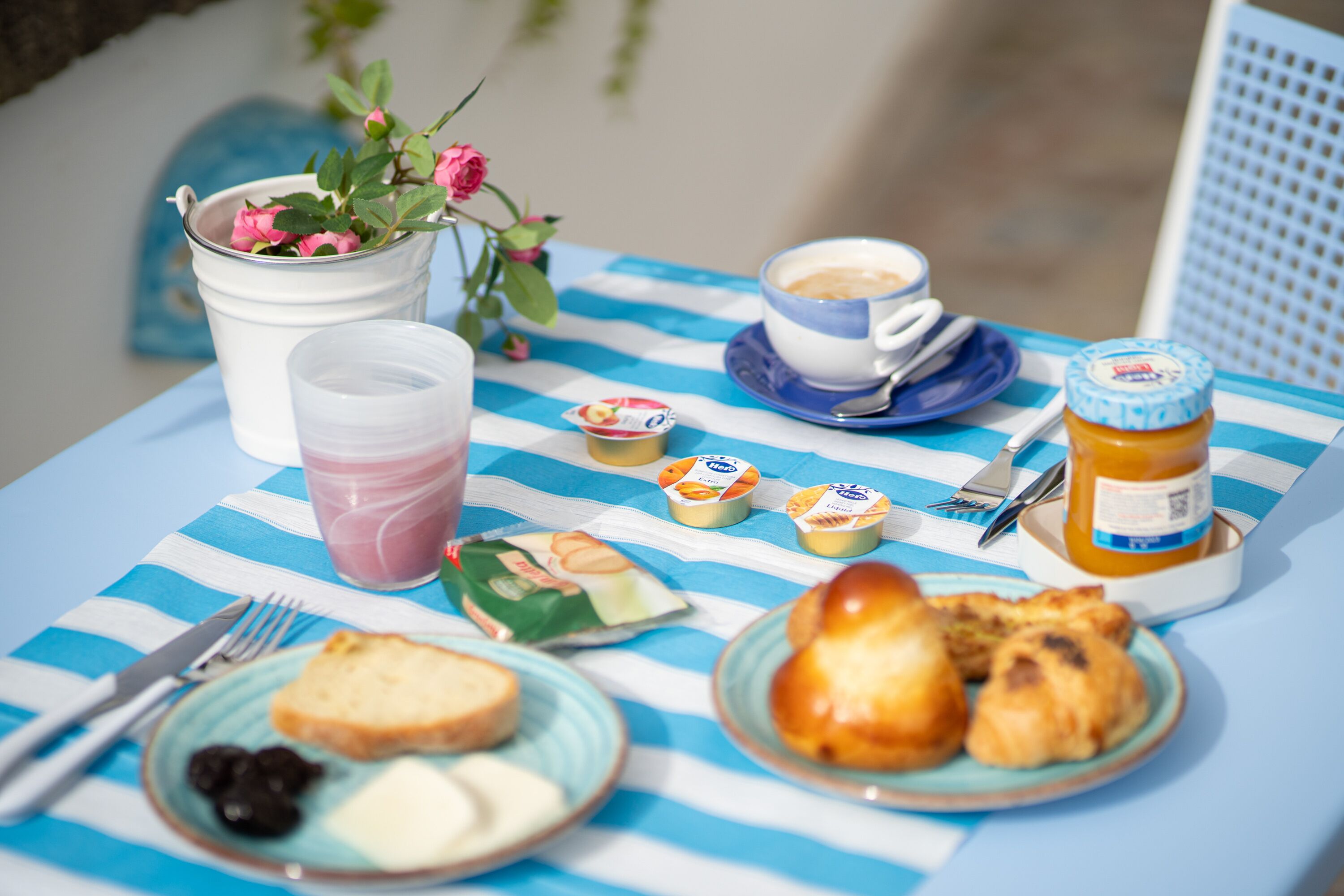 A table with a blue and white striped tablecloth, a cup of coffee, a jar of honey, a plate with pastries, a glass of juice, and a small flower arrangement.