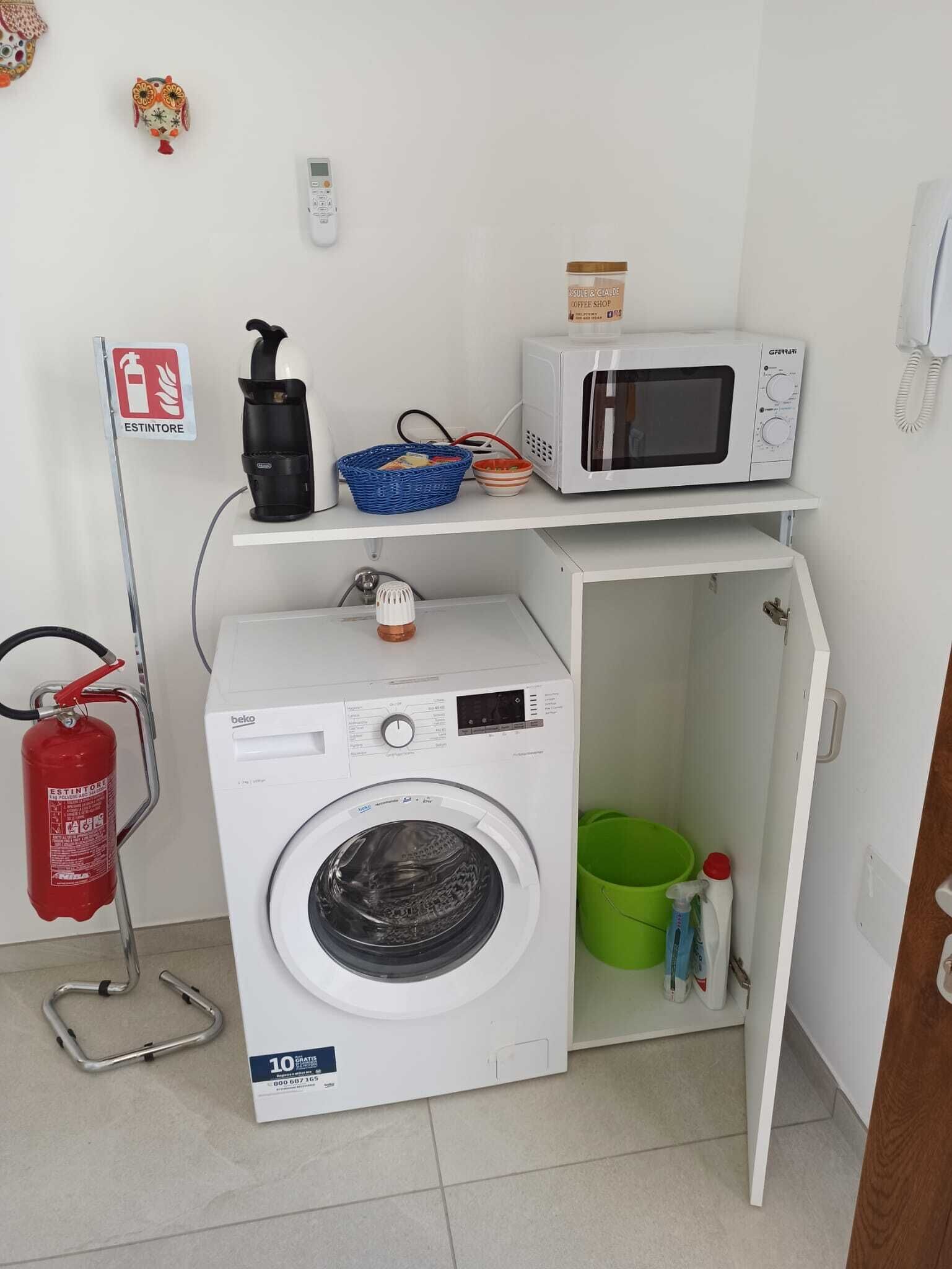 A compact laundry room with a washing machine, a microwave, and a small shelf with a basket and a coffee jar.