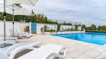 Une piscine bordée de chaises longues blanches et agrémentée d’un parasol blanc.