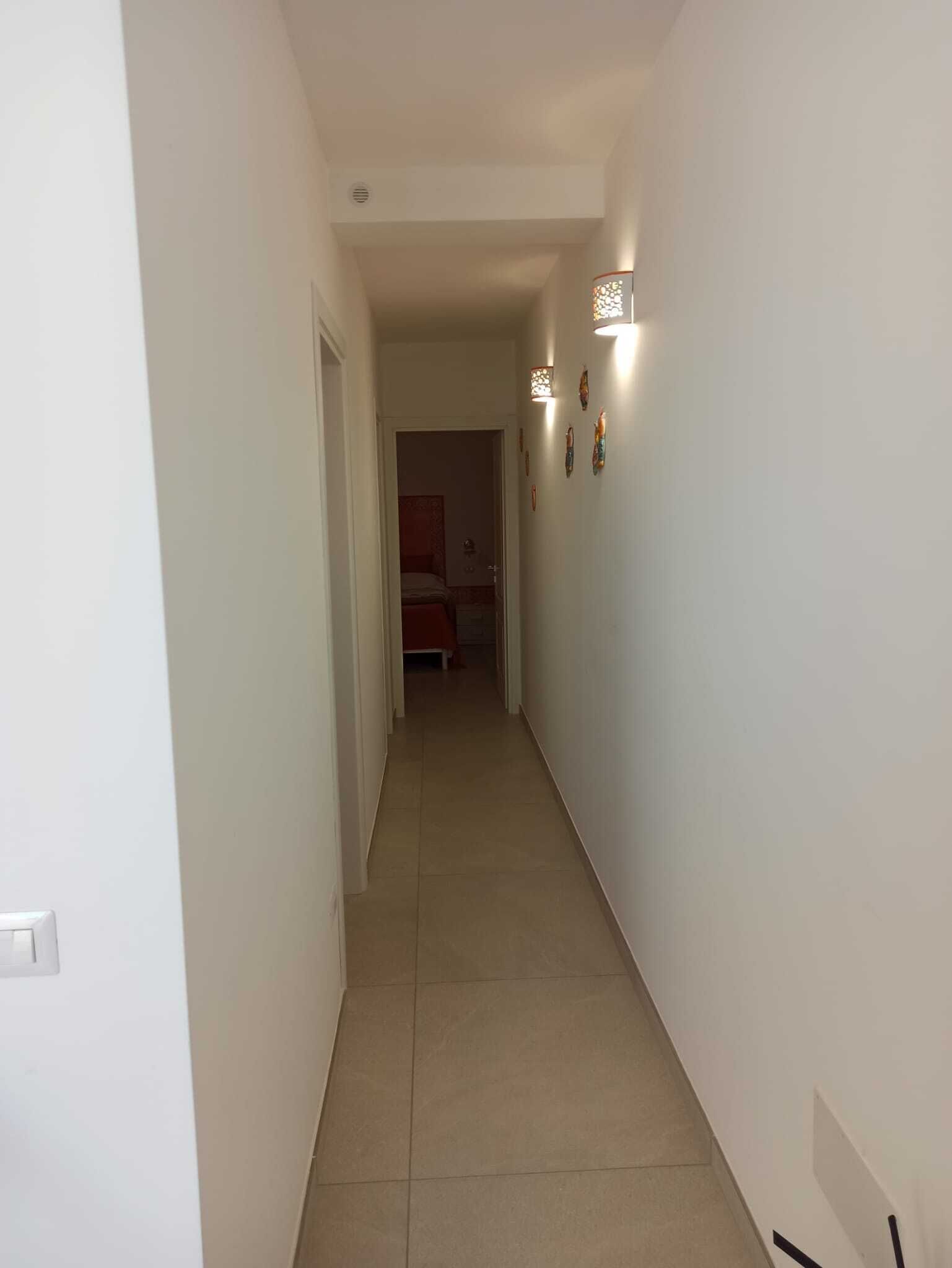 A narrow hallway with a door on the left and a bed in the room at the end.