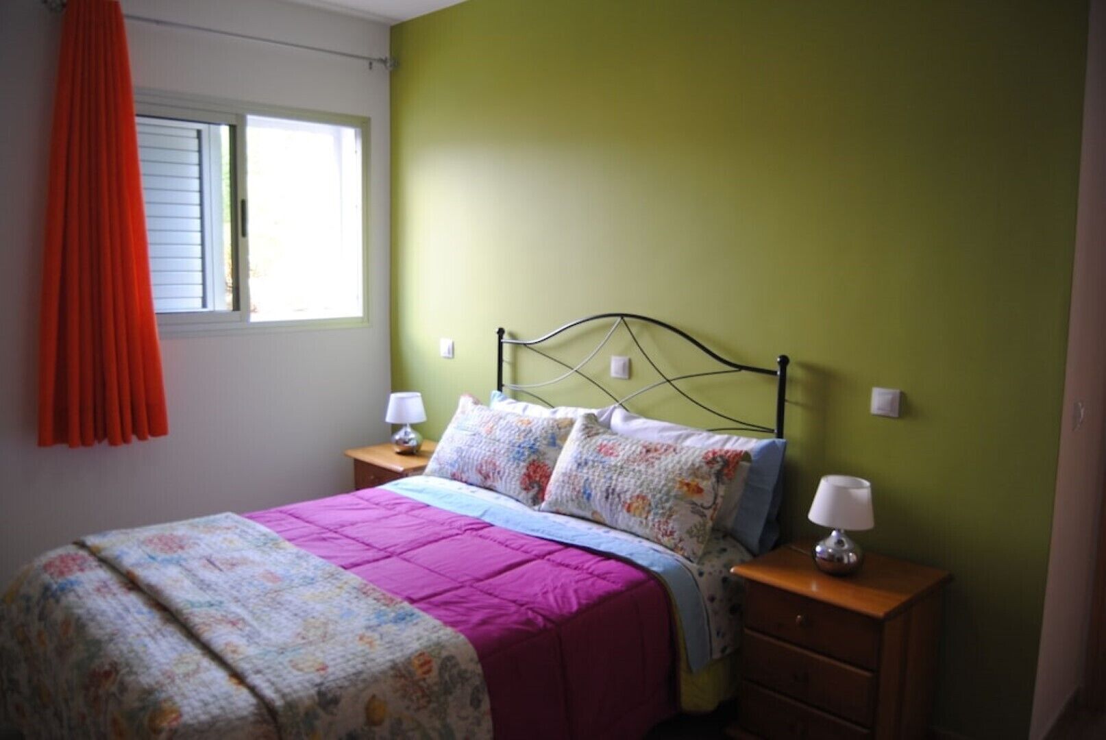Une chambre à coucher avec un lit, deux tables de chevet, une fenêtre avec des stores et un rideau.