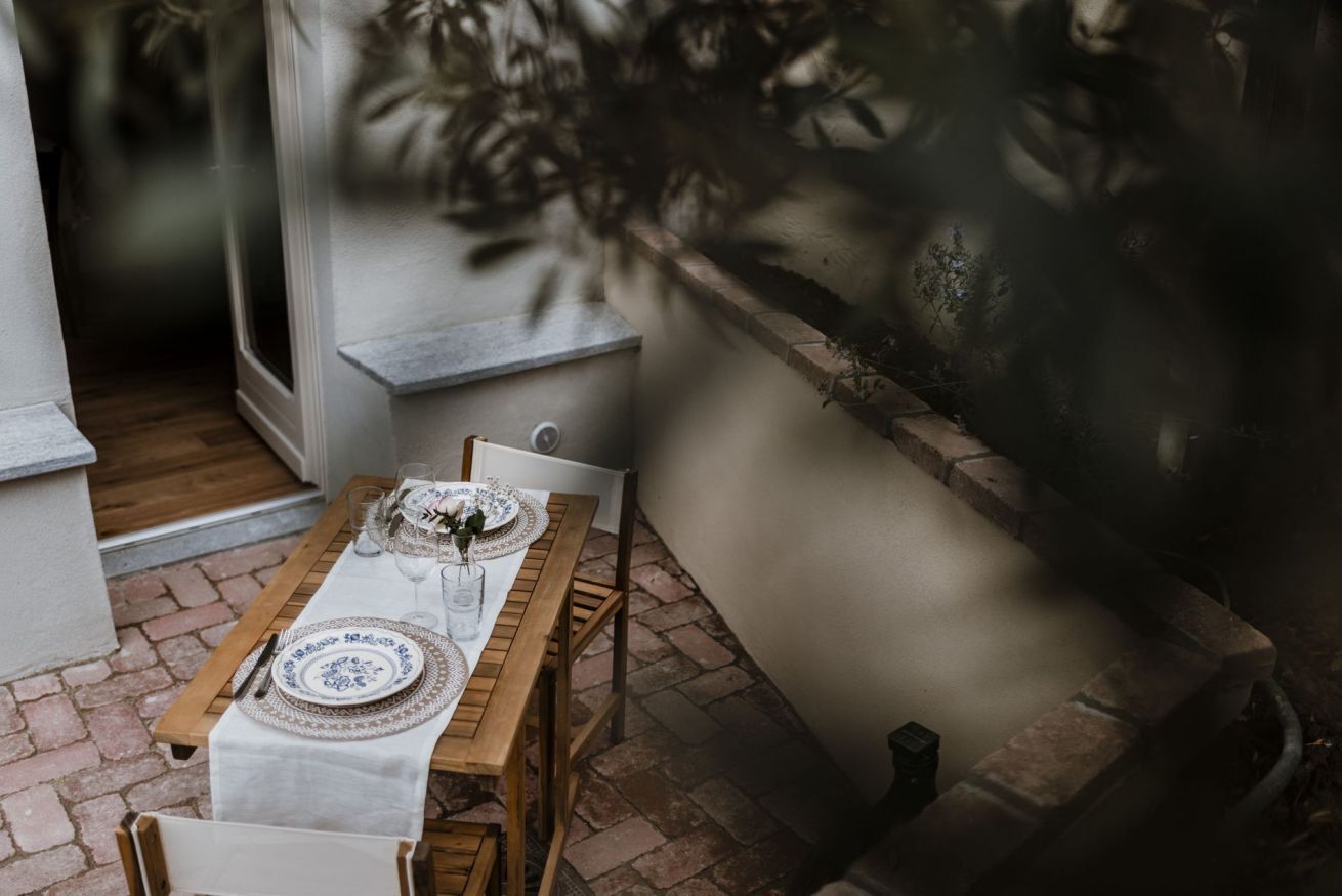 An outdoor dining area with a wooden table set for two, surrounded by brick flooring and a low stone wall.