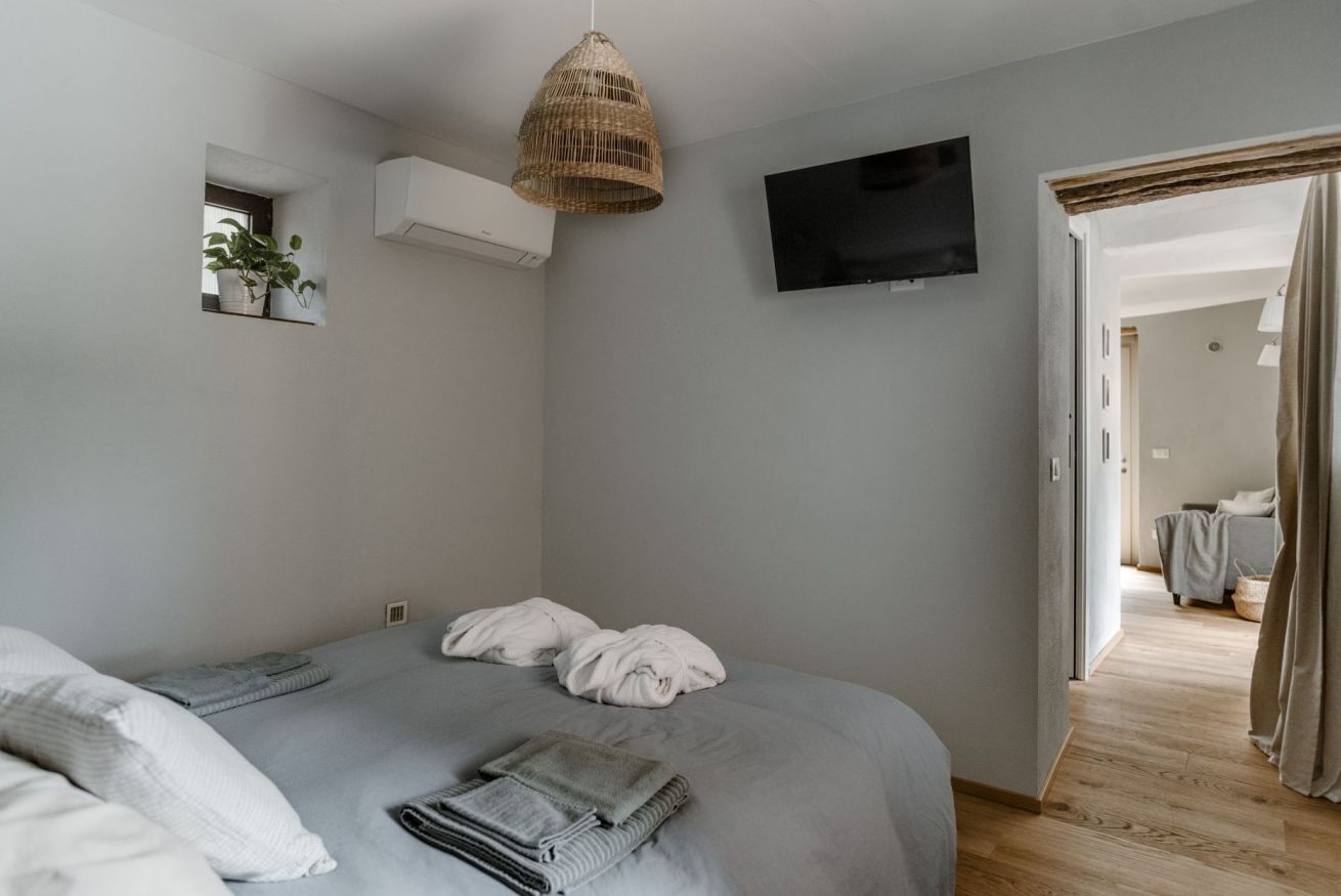 A bedroom with a bed, a television, a hanging light fixture, and a small window with a plant.