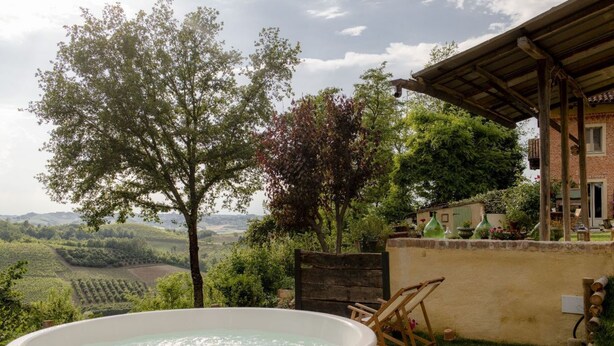 A hot tub in a garden with a view of a vineyard and a building with a wooden pergola.