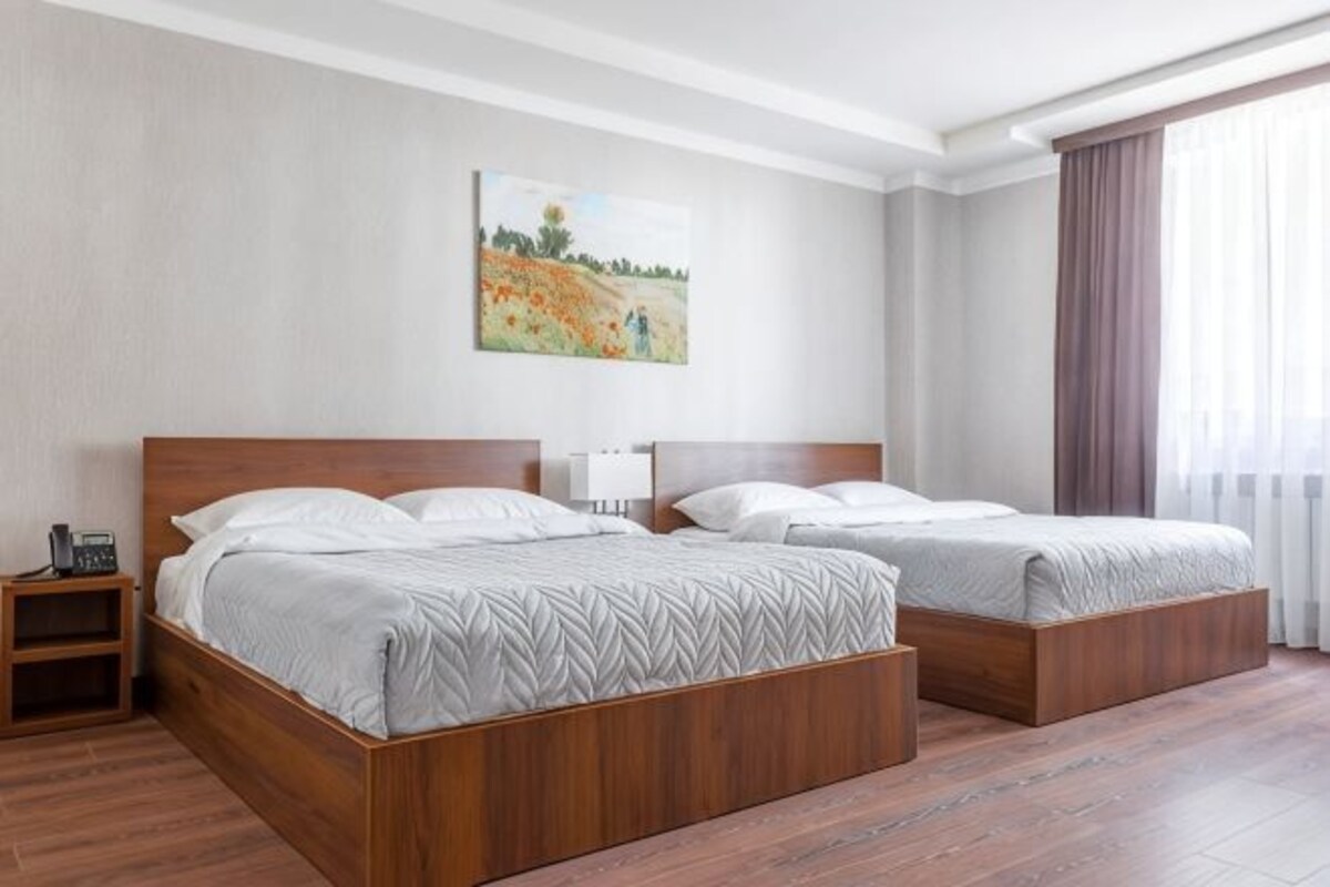 A hotel room with two beds, a nightstand, a painting on the wall, and a window with curtains.