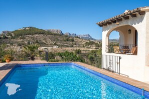 A swimming pool with clear blue water, surrounded by a white building with a tiled roof and an archway. In the background, there is a view of a hilly landscape with a building on top. - CostaBlancaDreams - Casa Narciso in Calpe (Calpe)