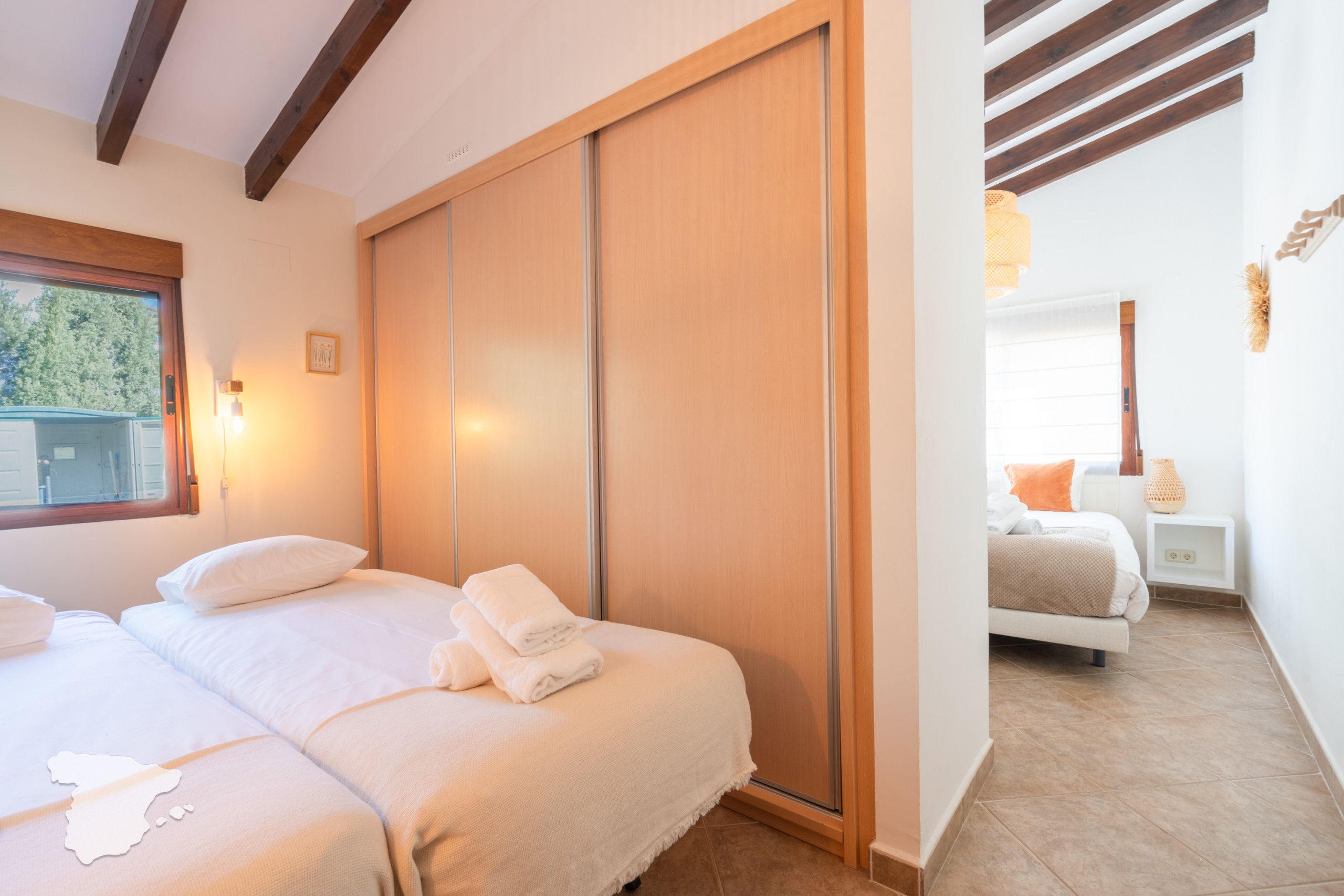 A bedroom with two beds, a sliding wooden door, a window with a view, and a bedside table with a lamp.