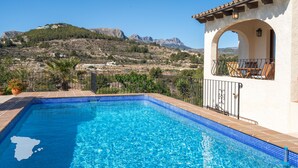 Una piscina con agua azul cristalina, rodeada por un edificio blanco con techo de tejas y un arco. Al fondo, se ve un paisaje montañoso con una edificación en la cima.