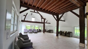 Amplia habitación con techos altos y vigas de madera a la vista, con varias zonas de estar con mobiliario de mimbre y grandes ventanales que ofrecen vistas al exterior.
