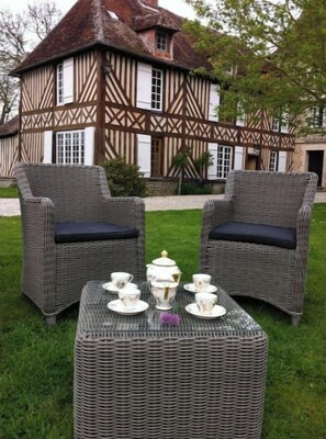 Il s’agit d’un bâtiment traditionnel de style Tudor, doté d’un toit de chaume et d’une structure en bois apparent. On y trouve, à l’avant, une table en osier sur laquelle est disposé un service à thé blanc, ainsi que deux chaises en osier avec des coussins foncés.