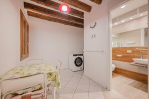 A small, clean room with a bed, a washing machine, and a bathroom with a sink and mirror. - Rialto Suite (Venice)