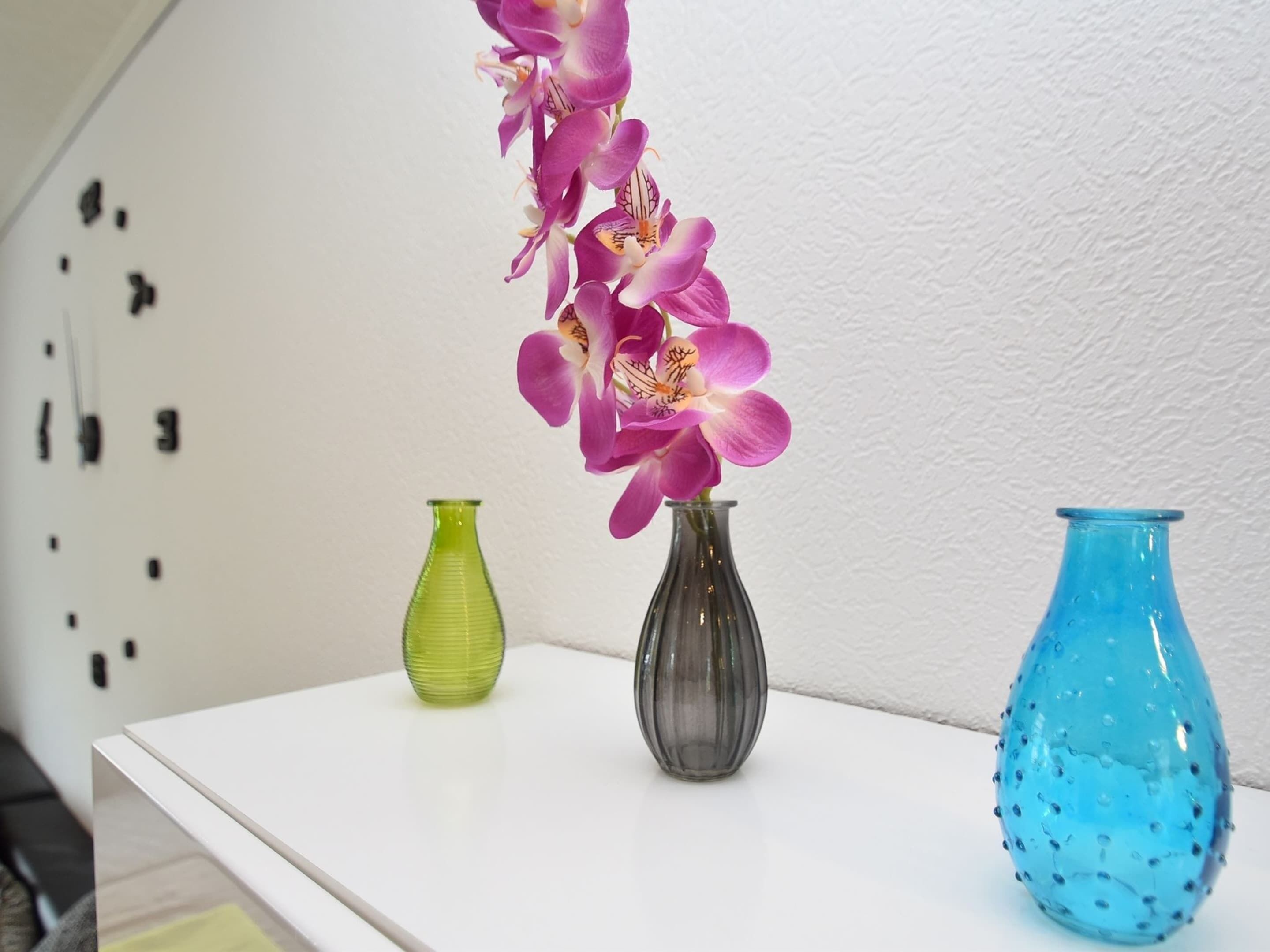 Três vasos de vidro sobre uma mesa branca, em frente a uma parede texturizada.