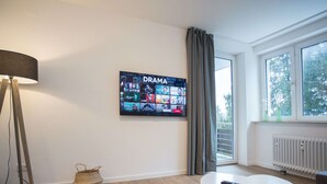 Un salon moderne avec un téléviseur à écran plat diffusant une chaîne de séries dramatiques, une table ronde blanche avec une télécommande, un lampadaire et un panier en osier.