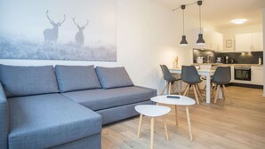 Un salon moderne avec un canapé d’angle gris, une petite table blanche et un coin repas composé de chaises, avec une cuisine en arrière-plan.