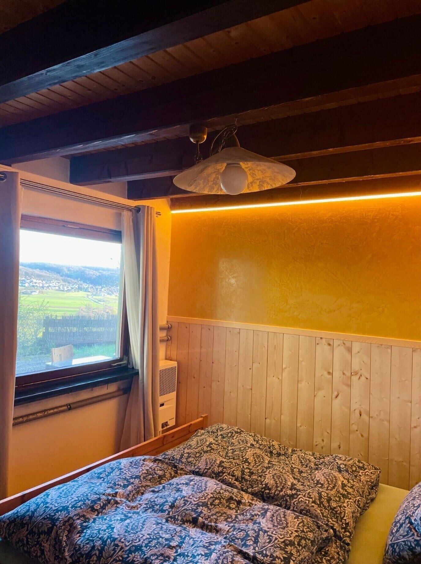 Una habitación con cama, ventana con cortinas, lámpara colgante y un calentador de pared.