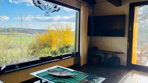 A room with a large window, a flat-screen TV, a table with a glass top, and a view of the countryside. - Ferienhaus, Schontal-winzenhofenn (Schöntal)