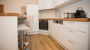 Una cocina compacta con armarios blancos, encimera de madera, horno empotrado, cafetera y un taburete de bar.