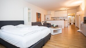 Un dormitorio moderno con una cama grande, un pequeño sofá, una mesita de centro y una zona de cocina al fondo.