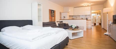 Un dormitorio moderno con una cama grande, un pequeño sofá, una mesita de centro y una zona de cocina al fondo.