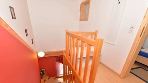 Un escalier en bois, doté d’une rampe, mène à une pièce où se trouvent un lit et une petite fenêtre.