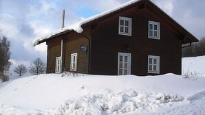 Une maison en bois avec un toit en pente recouvert de neige, entourée d’un paysage enneigé.