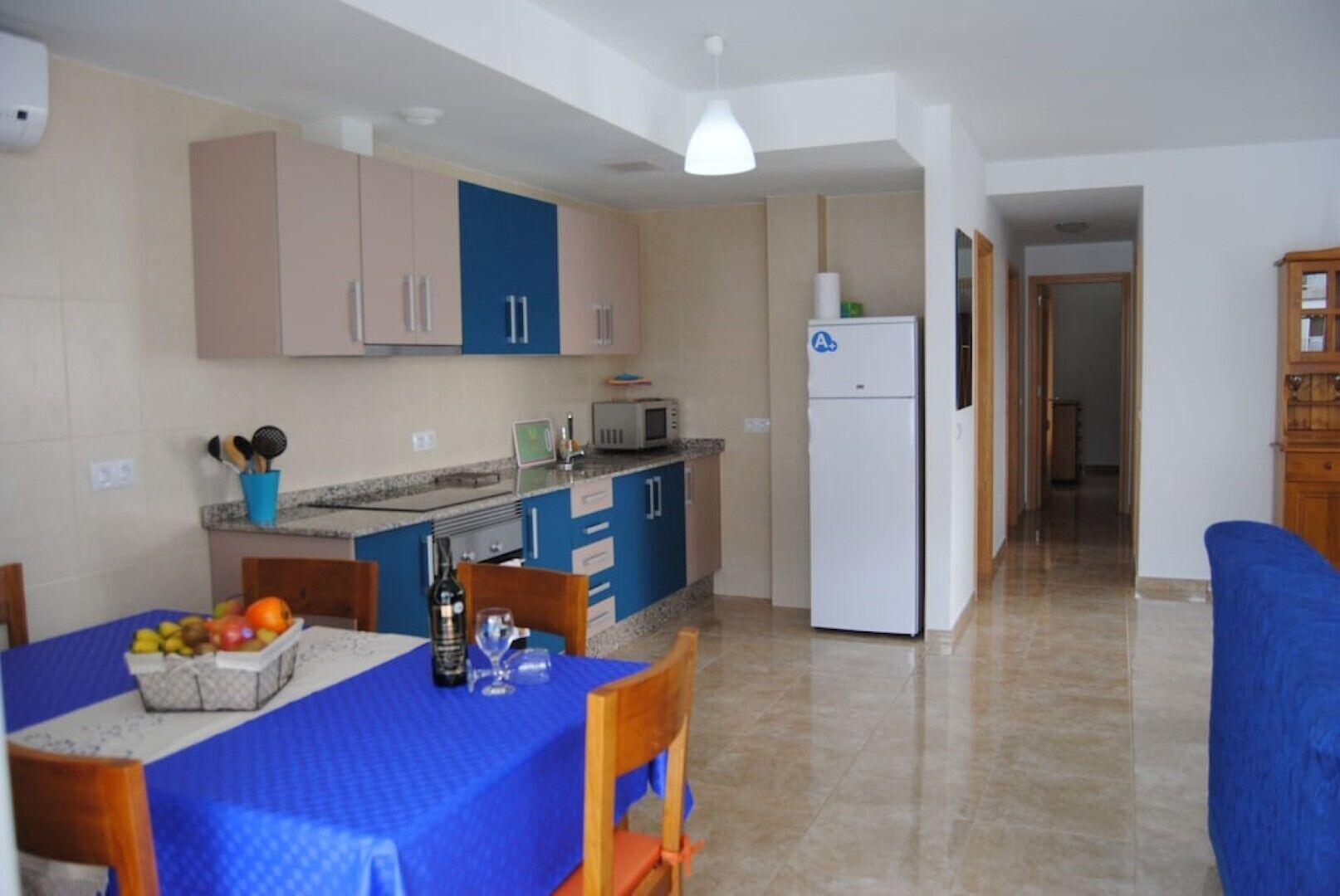 A compact kitchen with a dining area, featuring a refrigerator, microwave, and a dining table set with fruits and a bottle.