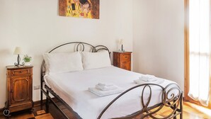 Cama bem arrumada com lençóis brancos e duas toalhas dobradas, mesa de cabeceira de madeira com abajur, e um quadro acima da cama.