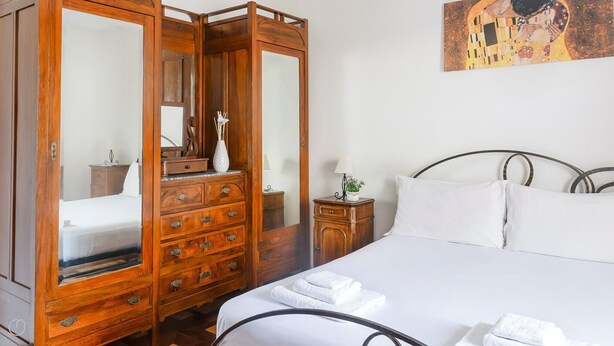 Quarto com guarda-roupa de madeira, cama com roupa de cama branca e pequena mesa de cabeceira com abajur.