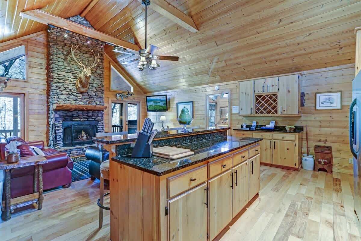 A log cabin interior with a stone fireplace, wooden cabinets, a kitchen island, and a leather sofa.