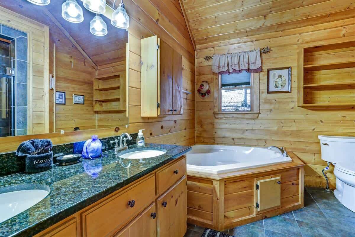 A wooden cabin bathroom with a large mirror, two sinks, a vanity, a bathtub, and a toilet.