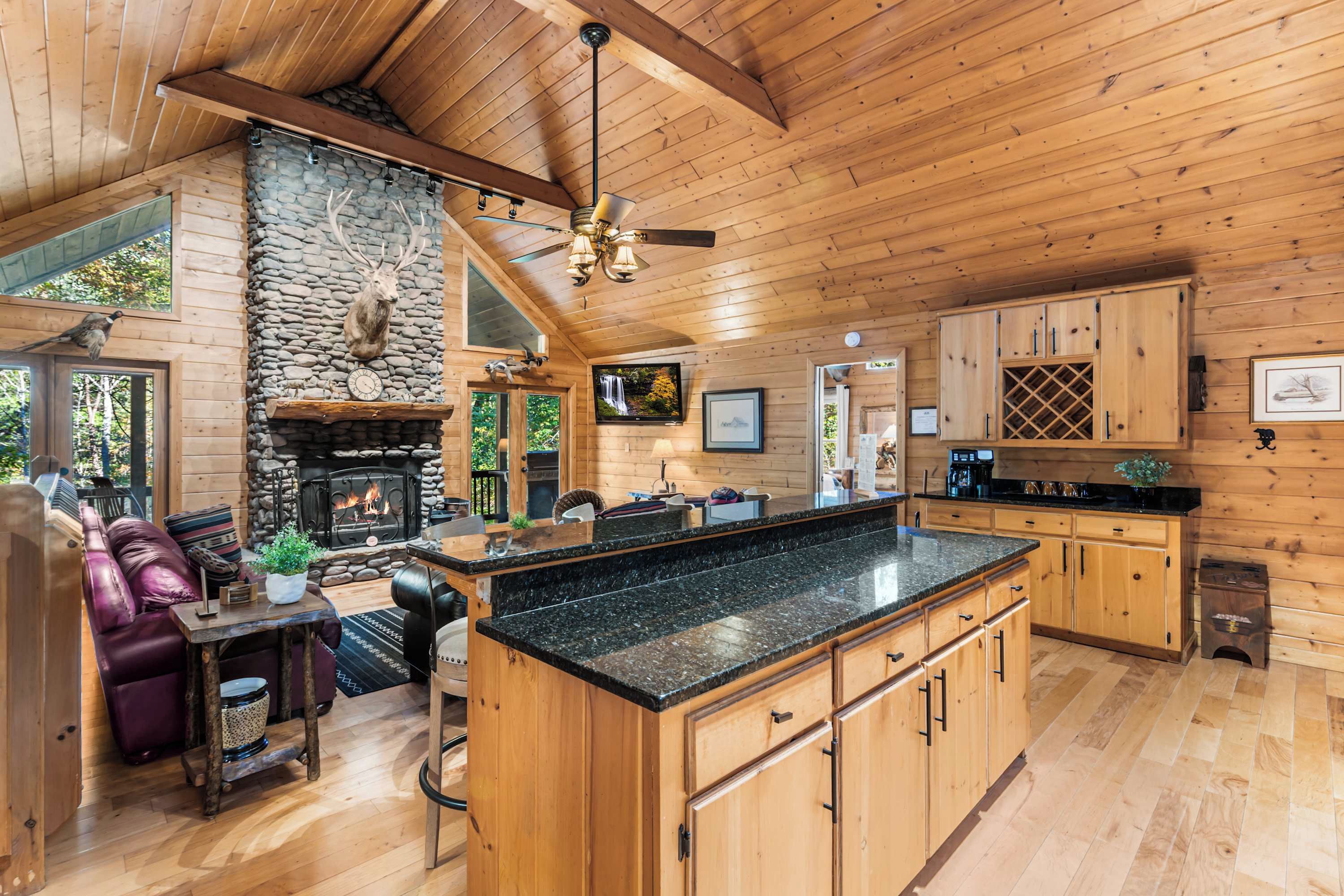 A spacious cabin interior with a stone fireplace, wooden ceiling, and a kitchen area with wooden cabinets.