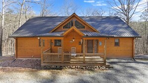 Une cabane en rondins avec un porche à l’avant, une cheminée et une grande fenêtre.
