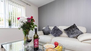 Een moderne woonkamer met een glazen salontafel, een witte bank met grijze kussens, een vaas met bloemen, wijnglazen en een schaal met fruit.