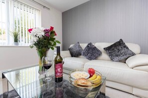 Una sala moderna con una mesa de centro de vidrio, un sofá blanco con almohadones grises, un jarrón con flores, copas de vino y un cuenco de frutas.