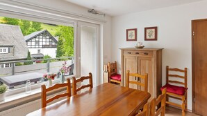 Un coin repas avec une table et des chaises en bois, un meuble contenant deux tableaux encadrés et une fenêtre donnant sur des maisons et de la verdure.