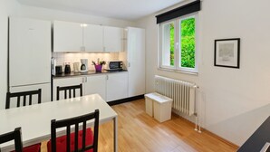 Une cuisine moderne avec des meubles blancs, une table à manger avec des chaises, une fenêtre donnant sur un espace verdoyant et un tableau encadré au mur.