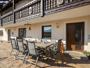 An outdoor dining area with a table and chairs, a wooden balcony with intricate railings, and a door with a glass panel. - Apartment in Gleissenberg With Garden & BBQ (Gleissenberg)