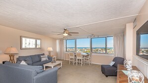 A living room with a sofa, a dining table with chairs, a ceiling fan, and large windows offering a city view. - Beautiful sunset and ocean view from 2 Bedroom Condo with Free WiFi and an Outdoor Pool Located in Midtown Only Steps to Beach! (Ocean City)