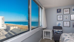 A room with a large window overlooking a beach and ocean, a gray armchair, a small white table, and several framed pictures on the wall. - Beautiful sunset and ocean view from 2 Bedroom Condo with Free WiFi and an Outdoor Pool Located in Midtown Only Steps to Beach! (Ocean City)