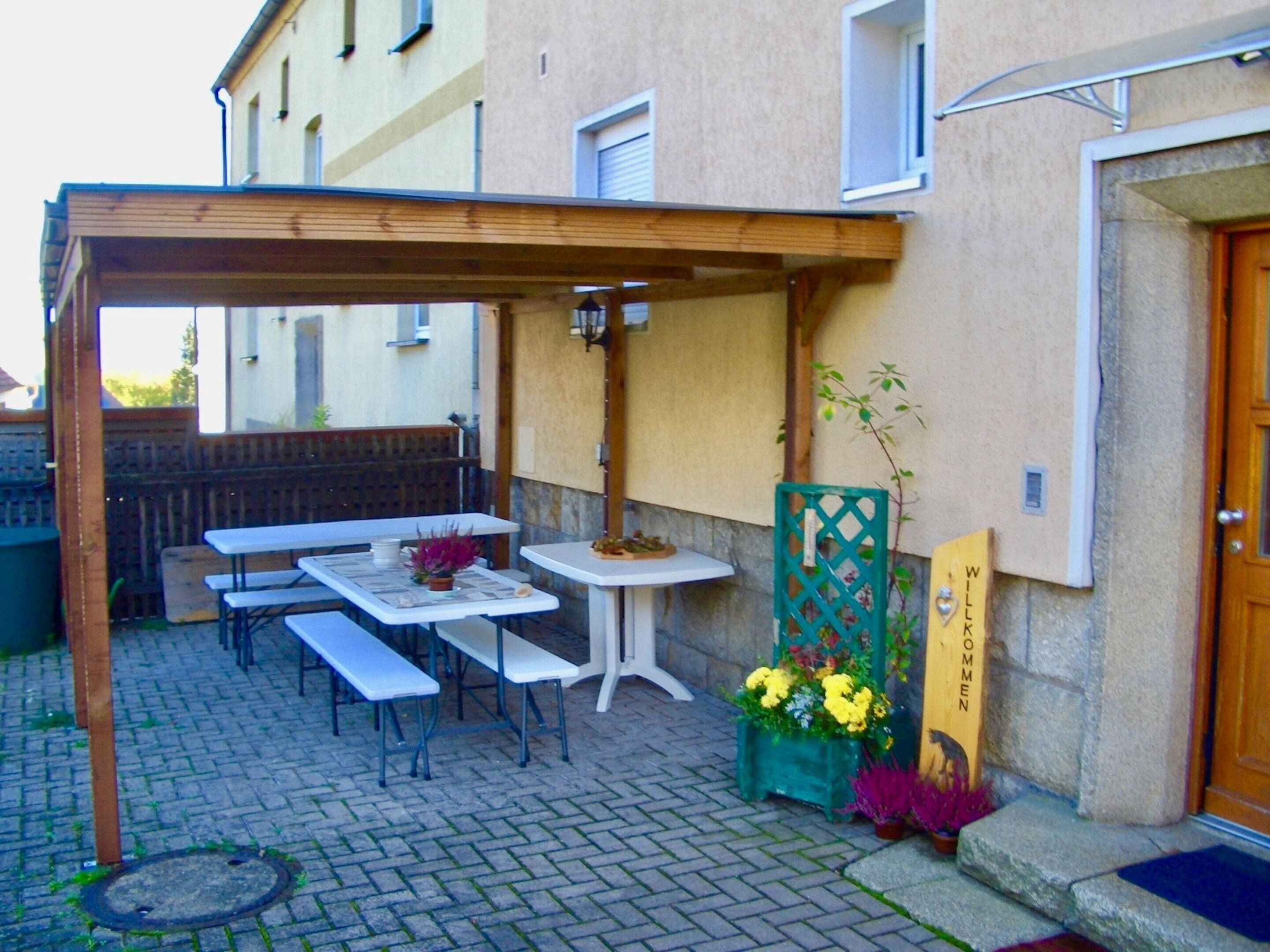 Un espacio de patio al aire libre con mesas, bancos, una pérgola de madera y un pequeño jardín con flores.