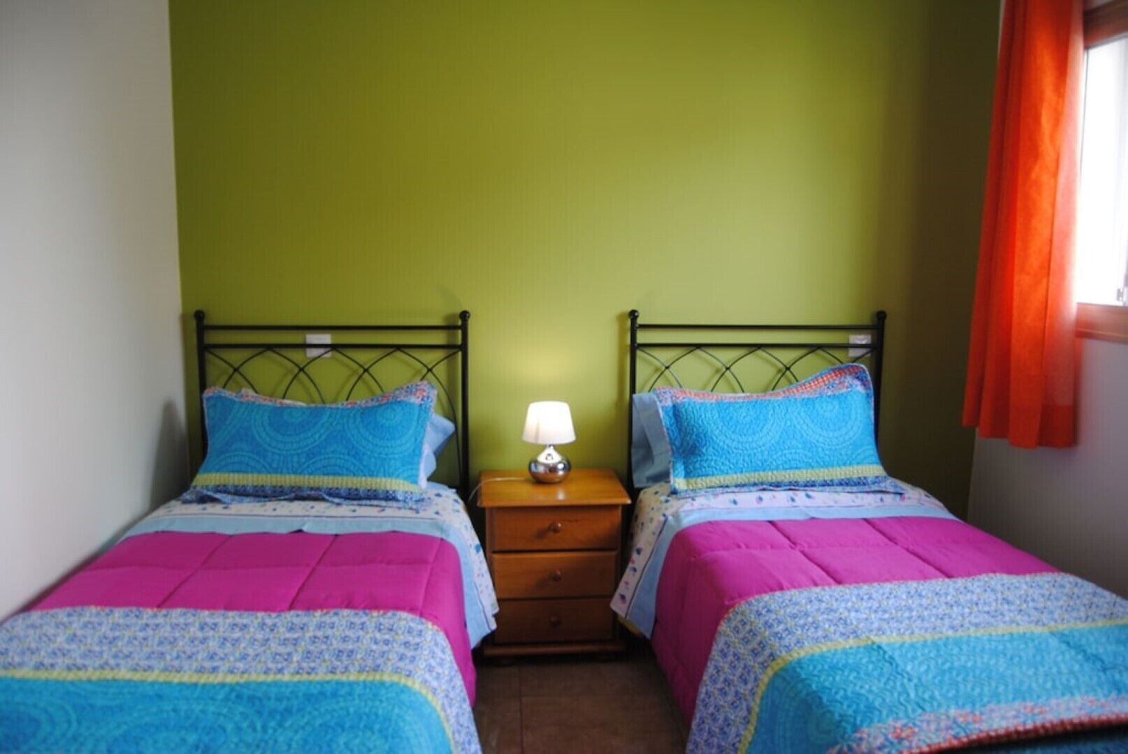 Una habitación con dos camas individuales, una mesita de noche con lámpara y una ventana con cortinas naranjas.