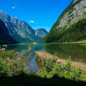 Um lago tranquilo com um píer de madeira, rodeado por montanhas verdes e céu azul límpido.