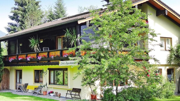 A two-story building with a balcony adorned with flowers, a sign reading "Joni's Apartment," and a small outdoor seating area. - Cosy Flat in Schonau am Konigssee (Schoenau am Koenigssee)