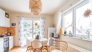 Une cuisine moderne avec des meubles blancs, une table à manger avec des chaises, un fauteuil en osier et une fenêtre donnant sur des arbres.