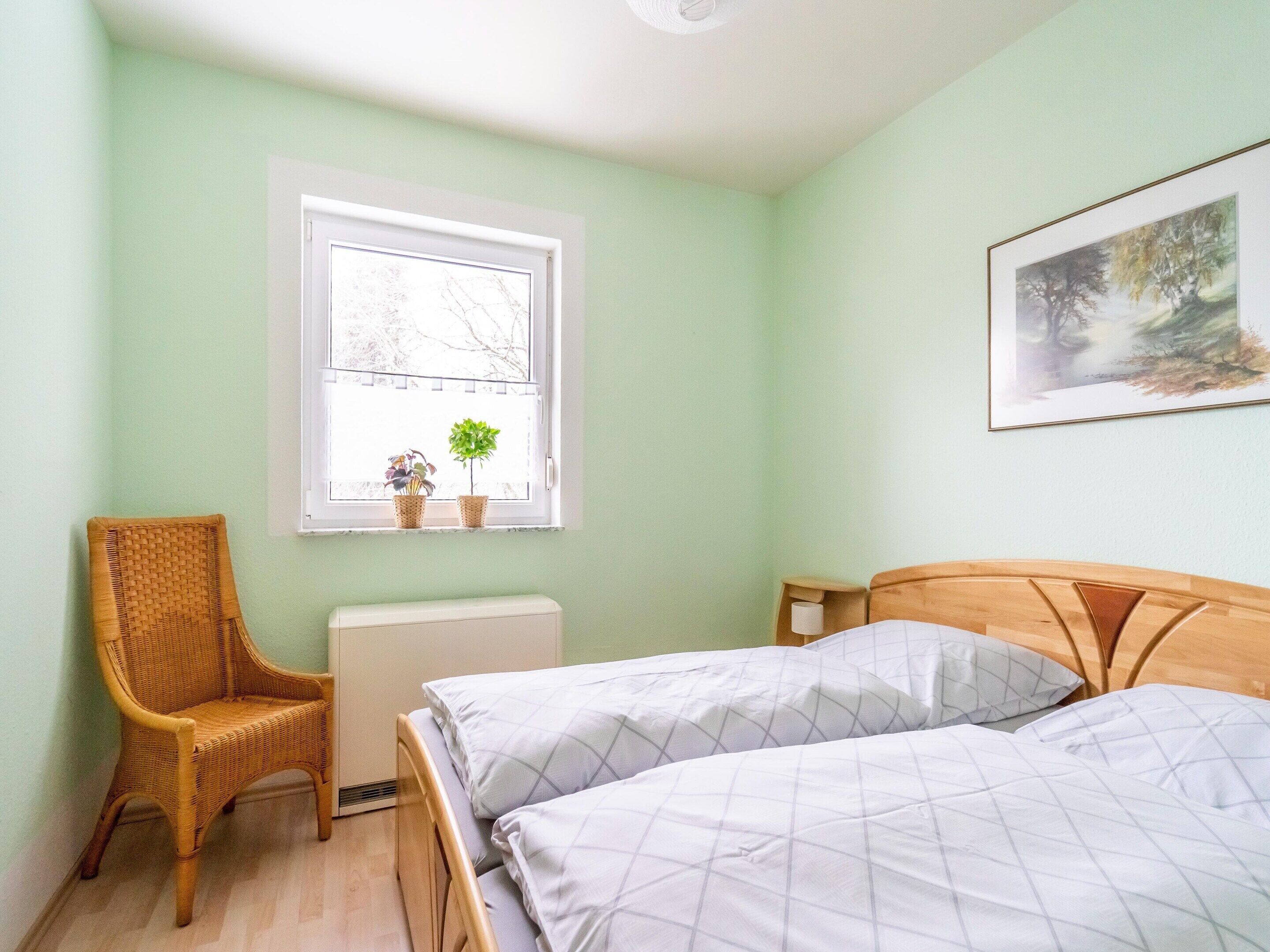 Een slaapkamer met twee bedden, een raam met uitzicht op bomen, een radiator, een rieten stoel en een schilderij aan de muur.