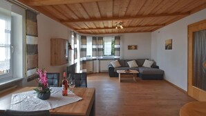 A spacious living area with a wooden ceiling, a sofa set, a dining table with chairs, and a view through large windows. - Apartment in the Bavarian Forest (Zenting)