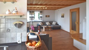 A cozy living room with a sofa, a coffee table, and a dining area with a basket of fruit. There is a kitchen area with a tiled backsplash and a statue on the wall. - Apartment in the Bavarian Forest (Zenting)