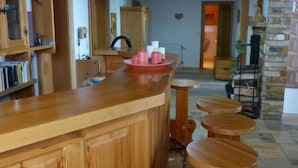 Un rincón con barra de madera y taburetes, una cocina al fondo y una chimenea de piedra.